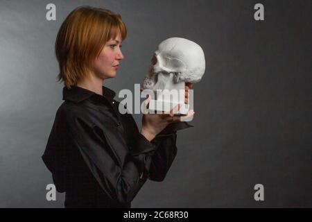 Carina giovane zenzero ragazza, in abito nero, tenendo un gesso cranio umano, lo guarda Foto Stock