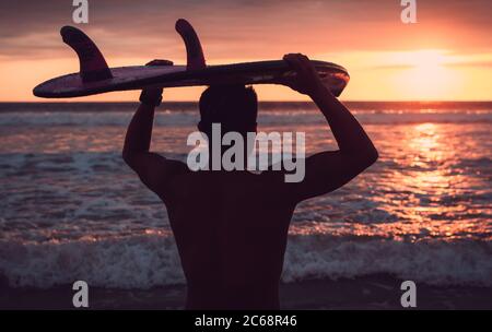 Retroilluminata di un surfista che tiene la sua tavola da surf sulla testa guardando il mare durante un incredibile tramonto nella spiaggia in Ecuador Foto Stock