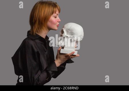 Carina giovane zenzero ragazza, in abito nero, tenendo un gesso cranio umano, lo guarda Foto Stock