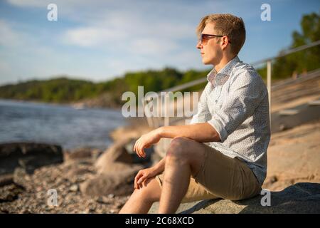 Pensieroso giovane uomo seduto su roccia guardando al mare Foto Stock