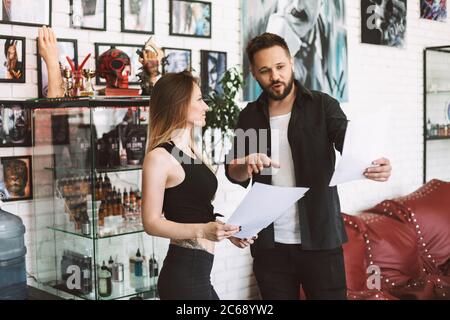 Tatuatrice e ragazza tatuata professionista che discute di nuovi schizzi tatuaggio passare il tempo in studio moderno Foto Stock