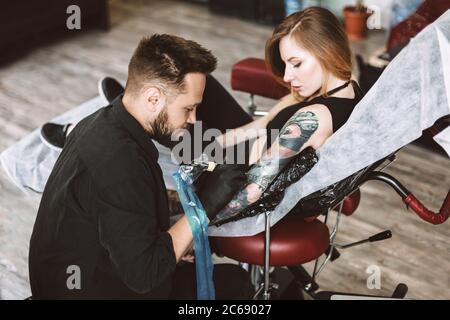 Tatuatore professionale che fa tatuaggio a mano da macchina tatuatrice mentre la ragazza guarda attentamente processo in studio Foto Stock
