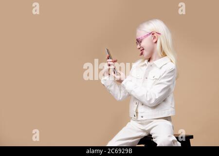 adorabile ragazza albino con aspetto affascinante scattare foto su telefono cellulare isolato, bambino prendere selfie, mostrare la lingua alla fotocamera, vista laterale Foto Stock