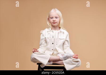 ritratto di adorabile bambino albino modello meditando in posa lotus, ragazza seduta con gambe incrociate, sguardo lato. albinismo concetto. mantenere la calma Foto Stock