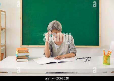 L'insegnante di senier teso stanco si tiene la testa con la mano mentre si siede dietro una paglia in classe. Foto Stock