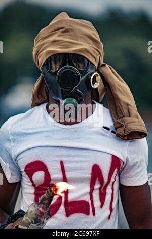Ritratto di nero afroamericano uomo in maschera a gas, ragazzo con iscrizione BLM sulla t-shirt è contro il razzismo e l'ingiustizia negli Stati Uniti, egli mette il fuoco su Foto Stock