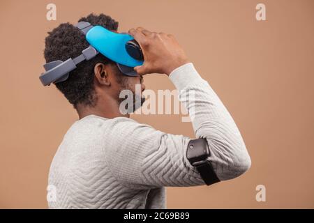 La realtà virtuale la tecnologia rende sempre grandi progressi. African casualmente vestito uomo cercando su occhiali VR isolate su sfondo beige Foto Stock