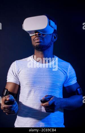 Di carnagione scura tourisistic maschio ventola con 3D tour in Asia, in piedi con head mounted display e controller palmare su sfondo scuro con rosso blu Foto Stock