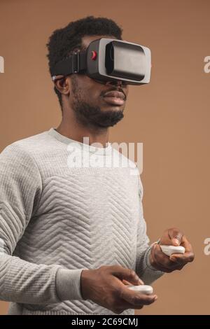 La realtà virtuale la tecnologia rende sempre grandi progressi. African casualmente vestito uomo cercando su occhiali VR isolate su sfondo beige Foto Stock