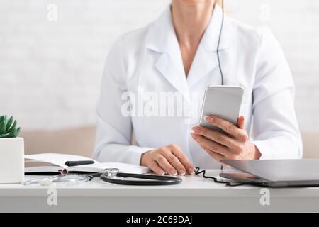 Visita al medico. Medico che guarda lo smartphone sul desktop, vicino sono stetoscopio blocco note e laptop Foto Stock