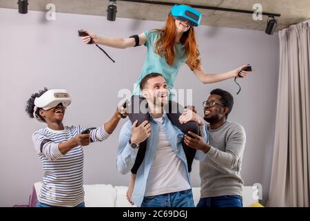 Gruppo multirazziale di persone divertendosi e dando piggyback ride per donna allegra in occhiali vr Foto Stock