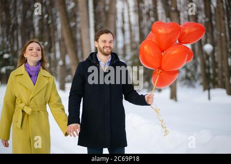 ritratto di giovane felice coppia, uomo e donna godere di camminare sulla strada invernale con palloncini d'aria rossa. bella coppia in cappotti celebrare san valentines Foto Stock