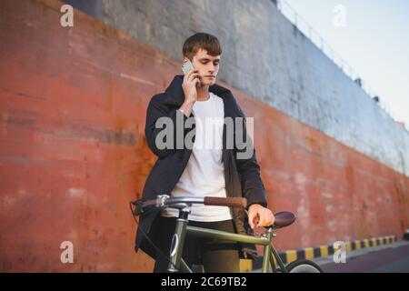 Giovane uomo con capelli marroni in piedi con bicicletta classica e guardando da parte pensieroso mentre parla sul suo cellulare. Ragazzo serio in giacca piumino e. Foto Stock