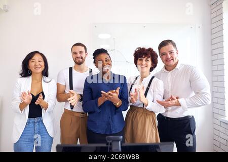 Sicuro sorridente gruppo multi-etnico loo alla macchina fotografica e applaudente, sfondo bianco lavagna a fogli mobili Foto Stock