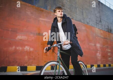 Ragazzo premuroso con i capelli marroni in bicicletta mentre sognando da parte. Giovane uomo in giacca e zaino nero in bicicletta sulla strada Foto Stock