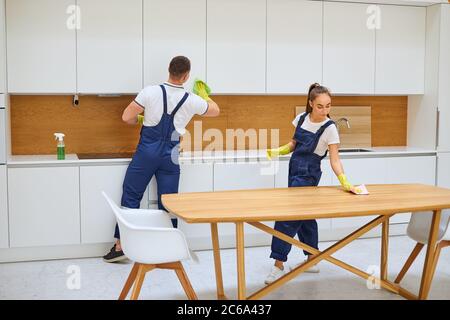 I giovani detergenti caucasici in cucina uniforme puliscono insieme la polvere dal tavolo di legno. Interno bianco Foto Stock