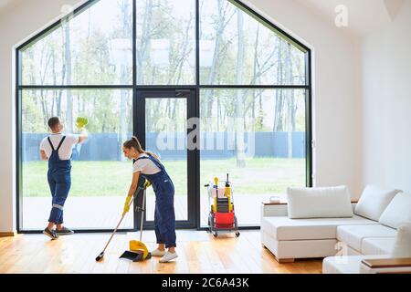 Vista posteriore su vigorosi gianti in uniformi blu che tengono le mop per lavare il pavimento e stracci per pulire i grandi vetri Foto Stock