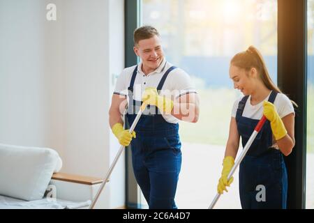 Il personale del servizio di pulizia lavora insieme, lavando il pavimento con la mop. Indossare indumenti speciali per la pulizia. Sfondo panoramico della finestra Foto Stock