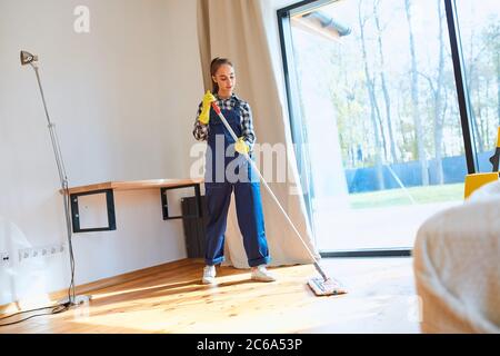 Perfetto lavoro di pulizia. Agnello attento in uniforme attentamente mop pavimento di legno in camera da letto, sfondo finestra. Foto Stock