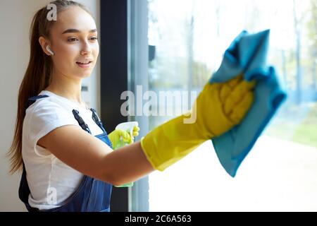 Giovane donna in uniforme, guanti di gomma gialla che reggono il panno blu per lavare il vetro, in cuffia Foto Stock