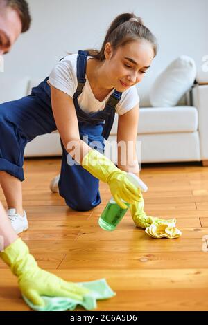 Giovane donna caucasica che indossa uniforme, utilizzando straccio e spray, lucido pavimento in legno, sfondo divano in pelle bianca Foto Stock