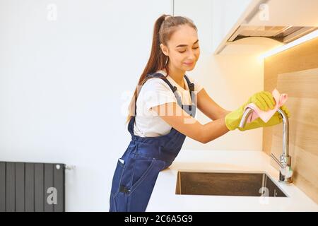 Giovane e bella ragazza janitor lavoro efficacemente nel servizio di pulizia, godere di lavoro. Donna che indossa guanti di gomma protettivi gialli, filo uniforme da lavoro Foto Stock