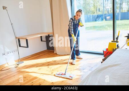 Perfetto lavoro di pulizia. Agnello attento in uniforme attentamente mop pavimento di legno in camera da letto, sfondo finestra. Foto Stock