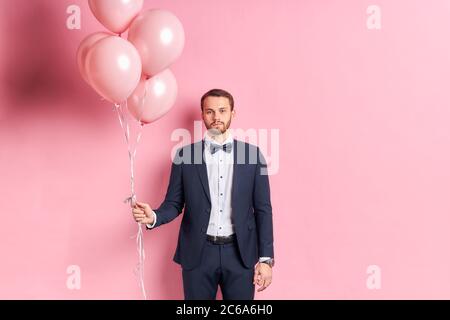 Triste maschio che indossa abito formale tenere palloncini rosa isolato su sfondo rosa. Emozioni e sensazioni tristi Foto Stock