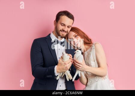 Sorridente coppia carina che indossa abiti da sposa e tiene i piccioni bianchi, stare felice isolato su sfondo rosa Foto Stock