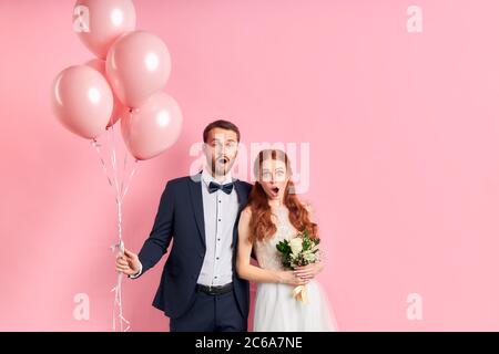 coppia di nozze che si posano insieme isolato su sfondo rosa. uomo in tuxedo che tiene balloni d'aria rosa, donna in bouquet bianco abito da sposa. Donna con Foto Stock