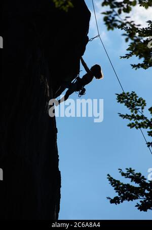 Silhouette di alpinista femminile arrampicata estremamente verticale roccia sotto cielo blu. Giovane donna che sale su masso alto e cerca di raggiungere la cima della montagna. Concetto di alpinismo, alpinismo, sport estremi. Foto Stock