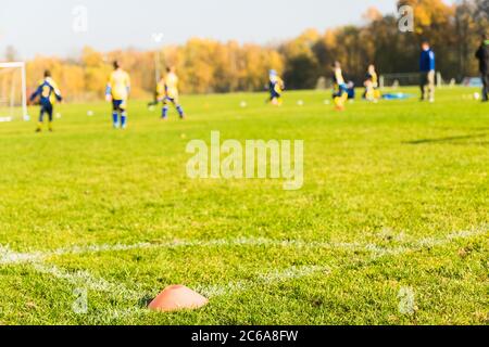 Fuoco selettivo per marchiare il cono in attrezzature di allenamento di calcio su verde con bambini poco nitidi giocatori di formazione sfondo. Foto Stock