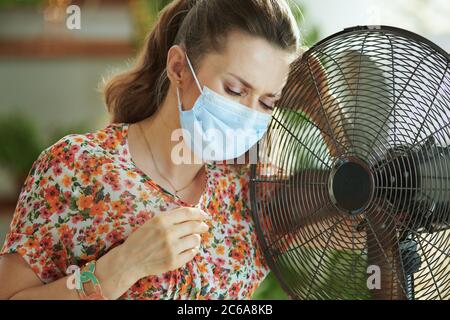 Estate calore. Infelice giovane donna in blusa floreale con ventilatore elettrico e maschera medica sensazione di calore estivo nella casa moderna in sole caldo giorno d'estate. Foto Stock