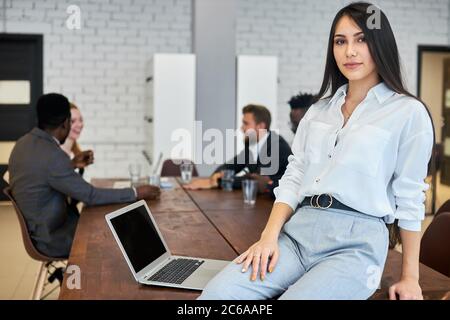 Donna sicura uomo d'affari, giovane e bella donna vestita di camicia bianca sedersi sul tavolo in posa e guardando la macchina fotografica Foto Stock