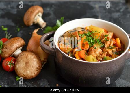 Stufate con pollo, patate, cipolle, funghi, pomodori e carote e prezzemolo. Stufato di verdure. Sfondo scuro. Foto Stock