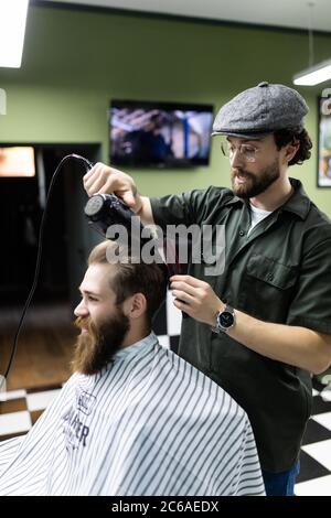 Già bello. Primo piano vista laterale di giovane uomo bearded che si cura dal parrucchiere con asciugacapelli al barbiere Foto Stock