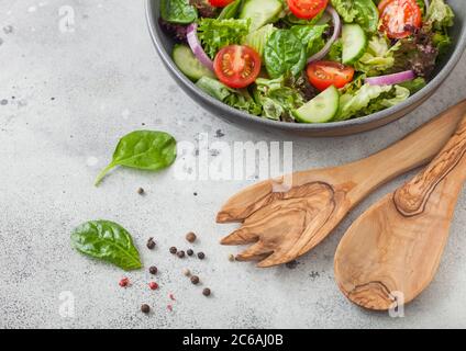 Ciotola grigia con insalata di verdure fresche sane con lattuga e pomodori, cipolla rossa e spinaci su sfondo chiaro del tavolo con cucchiaio di spatola e forchetta. Foto Stock