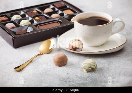 Scatola di selezione di caramelle al cioccolato di lusso con tazza di caffè nero e cucchiaio dorato su sfondo chiaro. Foto Stock