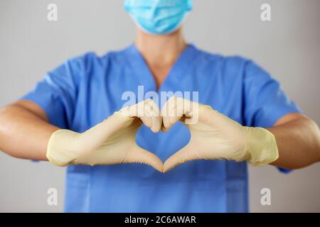 Medico in maschera e guanti che formano un cuore davanti a una camicia blu, Foto Stock
