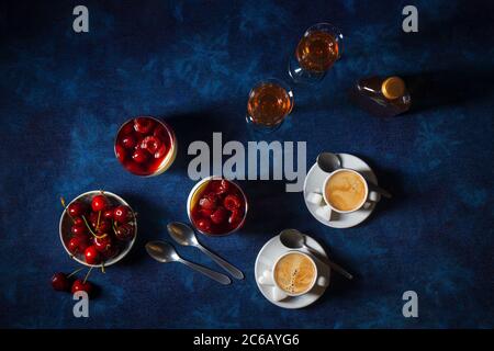 Panna cotta dessert tradizionale italiano con ciliegie in camicia al rum e sciroppo di miele. Tavolo di fondo blu scuro, ciotola delle ciliegie fresche, 2 bicchieri Foto Stock