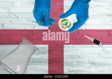 Bandiera inglese sul tavolo del laboratorio. Medico tecnico sanitario che tiene COVID-19 kit di raccolta tampone, indossando guanti di protezione blu, confe epidemico Foto Stock