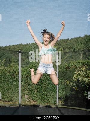 Foto verticale di un giovane bruna sorridente che salta su un trampolino con rete intorno al cortile verde all'aperto. La ragazza indossa jeans corti e. Foto Stock