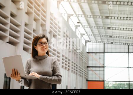 Ritratto di una donna esecutiva positiva che tiene un computer portatile in piedi in ufficio. Donna d'affari sorridente in casual in ufficio. Foto Stock