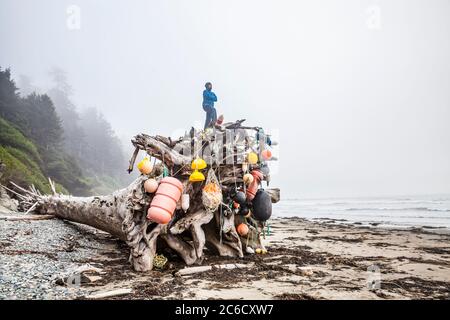 Un uomo si erge sulla cima di una gigantesca palla di radice di albero di driftwood decorata con boe lavate sulla Washington Coast, l'Olympic National Park Coastal Strip. Foto Stock