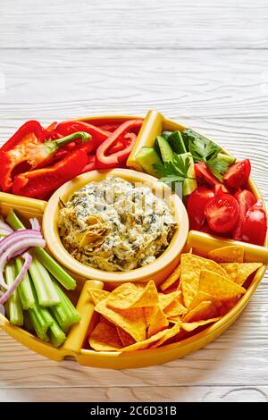 Set per immersione con formaggio di carciofi spinaci con patatine di tortilla, cetriolo, bastoncini di sedano, fette di pomodoro e peperone rosso, vista verticale dall'alto, primo piano Foto Stock