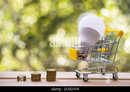 Pile di monete e lampadina a risparmio energetico nel mini carrello, sfondo verde naturale sfocato. Concetto di risparmio energetico e ambientale. Foto Stock