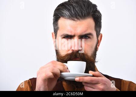 Il barman con il viso serio puzza di caffè. Concetto di ristorazione. L'uomo con barba e baffi trattiene una tazza di caffè su sfondo bianco. Il cameriere con la tazza di caffè bianca in mano si prepara a bere. Foto Stock