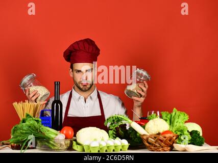 Chef con viso serio tiene vasi con porridge su sfondo rosso. Cuoco lavori  in cucina. Cucina e concetto di cucina. L'uomo in cappellino e grembiule si  pone con groat Foto stock 