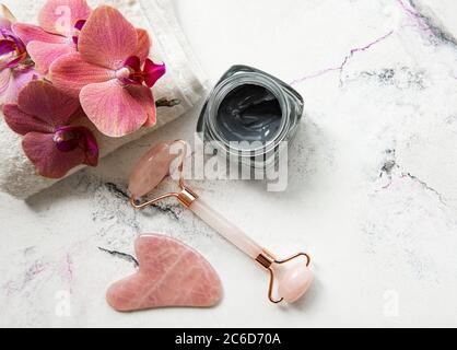 Cura della pelle mattina routine di bellezza a casa bagno - viso jade rullo massaggiatore per benessere terapia cosmetica, maschera e fiori orchidee rosa su marmo bac Foto Stock