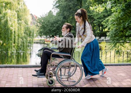 Bella ragazza hipster con capelli dreadlock che spinge la sedia a rotelle con l'uomo anziano, divertirsi nel parco della città di fronte al lago. L'uomo handicappato è Foto Stock